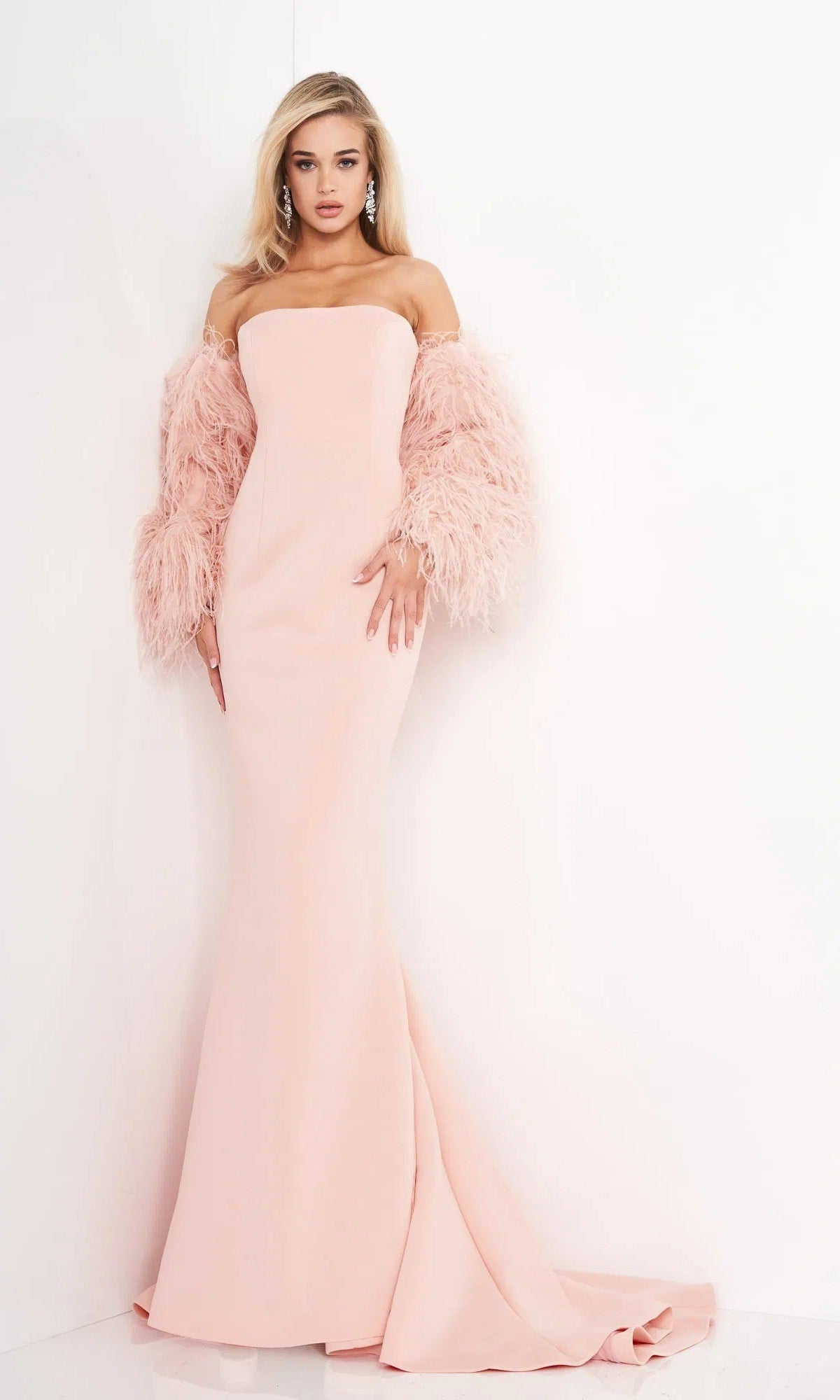 A woman stands against a white wall wearing the Jovani 1226 long prom dress in blush pink, featuring dramatic feather sleeves and accessorized with drop earrings.